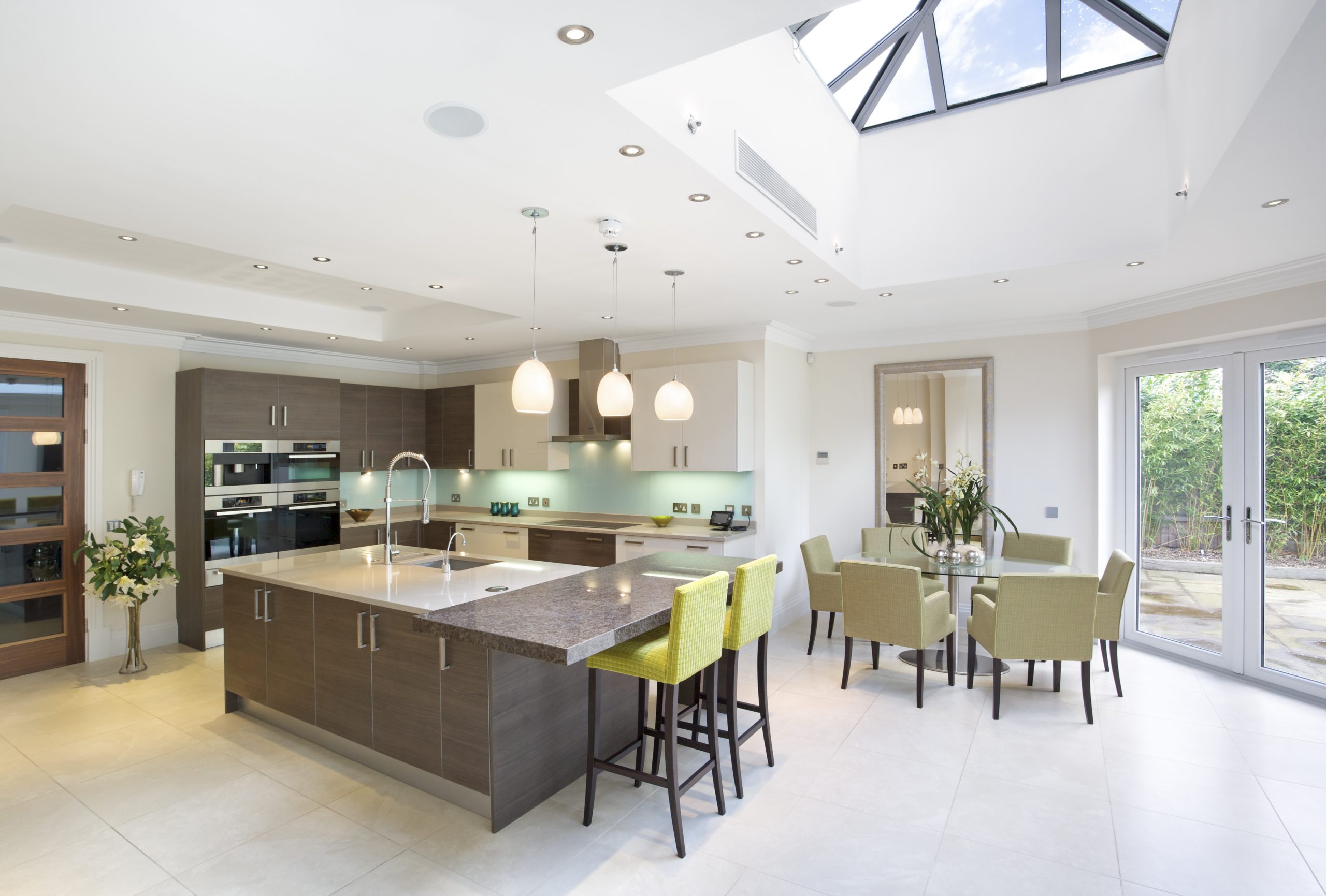 a large kitchen with light dining area in an expensive new home. An island at the centre of the kitchen has a breakfast bar attached and a sink with a large extendable tap and a "boiling-water-on-demand" tap with filtered water. Beneath a large skylight is a small table with six chairs. The kitchen decor is two toned with walnut panelling and cream coloured tops and facias.